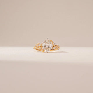 Front view of a oval diamond ring in 14KT Yellow Gold, featuring a floral vine band and sparkling accent diamonds against a beige backdrop.