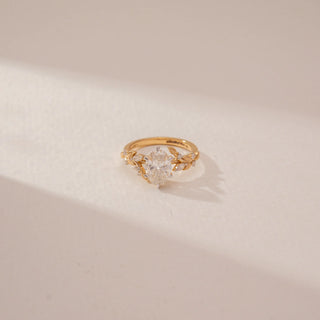 Close-up view of a oval diamond ring in 14KT Yellow Gold, featuring a floral vine band and sparkling accent diamonds against a beige backdrop.