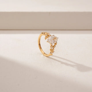 Angled view of a oval diamond ring in 14KT Yellow Gold, featuring a floral vine band and sparkling accent diamonds against a beige backdrop.
