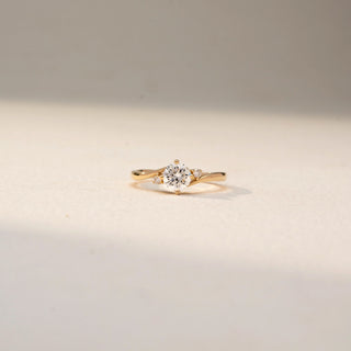 Front view of a solitaire diamond ring in 14 KT Yellow Gold, featuring a round center diamond accented by tapered side diamonds on a sleek crossover band against a soft beige backdrop.
