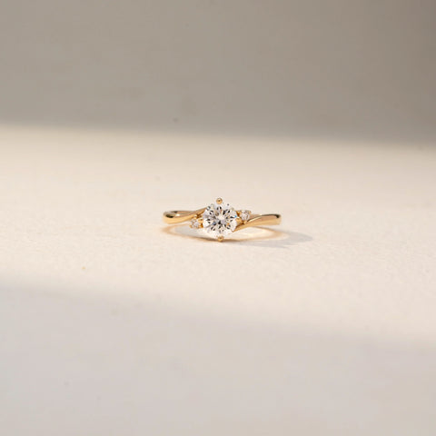 Front view of a solitaire diamond ring in 14 KT Yellow Gold, featuring a round center diamond accented by tapered side diamonds on a sleek crossover band against a soft beige backdrop.
