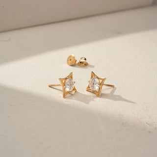 Angled view of geometric diamond stud earrings in yellow gold, featuring round brilliant-cut diamonds framed by sculptural openwork settings. Displayed on a beige backdrop, the design showcases modern elegance.