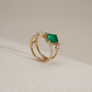 Side view of the Emerald Bloom Luxury Diamond Ring in 14KT Yellow Gold, featuring a vivid emerald-cut green centre stone accented with floral-inspired EF VVS diamonds on a diamond-studded band, displayed on a light textured surface.