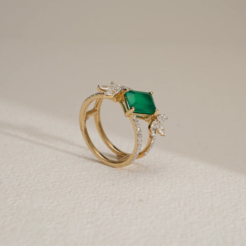 Side view of the Emerald Bloom Luxury Diamond Ring in 14KT Yellow Gold, featuring a vivid emerald-cut green centre stone accented with floral-inspired EF VVS diamonds on a diamond-studded band, displayed on a light textured surface.