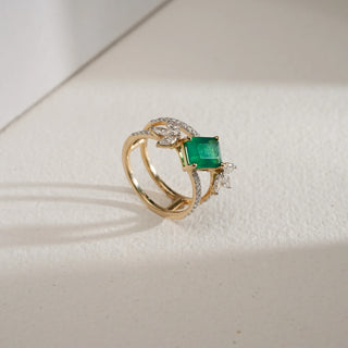 Angled view of the Emerald Bloom Luxury Diamond Ring in 14KT Yellow Gold, showcasing a vivid emerald-cut green gemstone with floral-inspired EF VVS diamond accents on a split diamond-studded band, placed on a light textured surface.