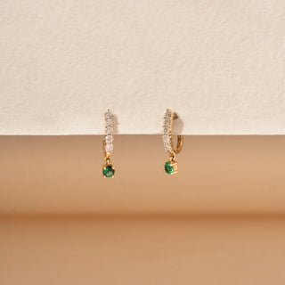 Close up view of round lab-grown diamond drop earrings in 14 KT white gold, featuring diamond huggie hoops with round emerald drop charms in a timeless elegant design