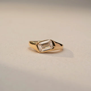 Front view of an emerald-cut solitaire signet ring in 14 KT Yellow Gold, featuring a bezel-set emerald-cut lab-grown diamond in a sleek tapered band, displayed against a beige backdrop.