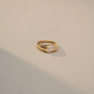 Side view of an emerald-cut solitaire signet ring in 14 KT Yellow Gold, featuring a bezel-set emerald-cut lab-grown diamond in a sleek tapered band, displayed against a beige backdrop.