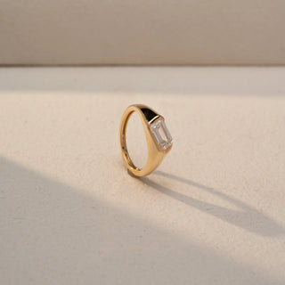 Angled view of an emerald-cut solitaire signet ring in 14 KT Yellow Gold, featuring a bezel-set emerald-cut lab-grown diamond in a sleek tapered band, displayed against a beige backdrop.