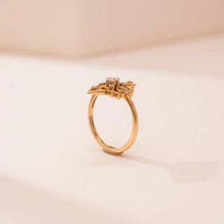 Side view of Reia’s 14 KT Yellow Gold Heritage Petal Ring. The diamond-set floral top is shown from the side on a thin gold band against a soft pink surface.