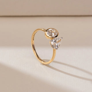 Side angle view of 14KT yellow gold open ring featuring an oval-cut diamond in a bezel setting and a pear-shaped diamond in a prong setting on a polished band.