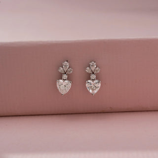 Front view of heart-shaped diamond drop earrings with pear-shaped and round accent stones, displayed against a soft pink backdrop for an elegant, romantic look.