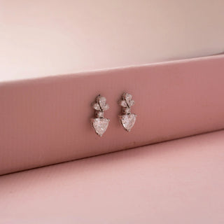 Side view of heart-shaped diamond drop earrings with pear-shaped and round accent stones, highlighting the raised setting, profile depth, and elegant craftsmanship on a pink backdrop.