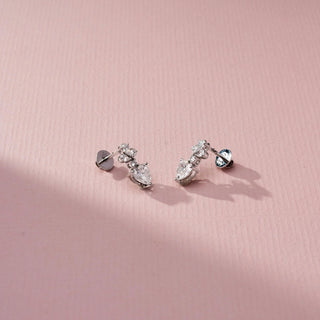 Angled view of heart-shaped diamond drop earrings with pear-shaped and round accent stones, highlighting the dimensional setting, post-back design, and sparkle on a soft pink surface.