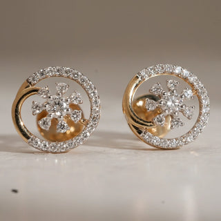 Close-up of floral diamond stud earrings in 14KT Yellow Gold, featuring a central diamond cluster framed by a circular diamond halo, placed on a neutral surface.
