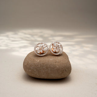 Front view of floral diamond stud earrings in 14 KT Rose Gold, featuring marquise-cut diamond petals within pavé-set circular frames, displayed on a textured stone surface.
