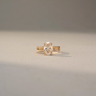 Front view of an oval solitaire engagement ring in 14 KT Yellow Gold, featuring an oval-cut lab-grown diamond in a four-prong setting with a pavé-set diamond band, displayed against a beige backdrop.