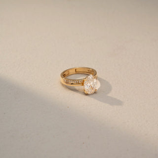 Side view of an oval solitaire engagement ring in 14 KT Yellow Gold, featuring an oval-cut lab-grown diamond in a four-prong setting with a pavé-set diamond band, displayed against a beige backdrop.