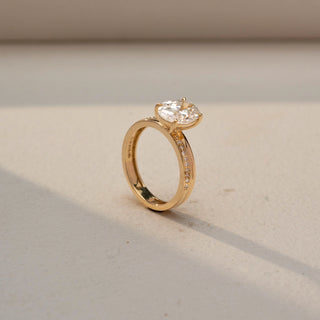 Angled view of an oval solitaire engagement ring in 14 KT Yellow Gold, featuring an oval-cut lab-grown diamond in a four-prong setting with a pavé-set diamond band, displayed against a beige backdrop.
