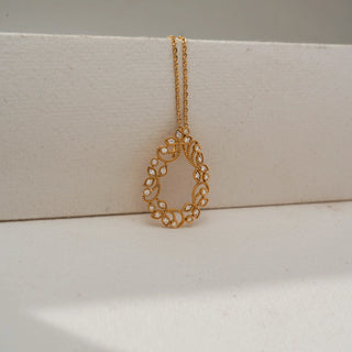 Close-up front view of a gold teardrop pendant necklace with intricate floral filigree and sparkling stone accents, displayed against a soft neutral background.