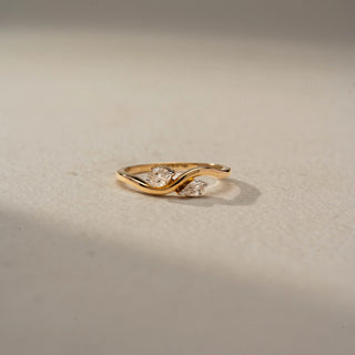 Front view of a bypass diamond ring in 14 KT Yellow Gold, featuring a round and a pear-shaped lab-grown diamond embraced by flowing sculpted gold curves in an elegant open design, displayed against a beige backdrop.