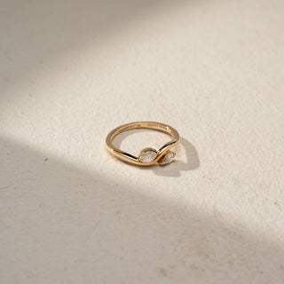 Top view of a bypass diamond ring in 14 KT Yellow Gold, featuring a round and a pear-shaped lab-grown diamond embraced by flowing sculpted gold curves in an elegant open design, displayed against a beige backdrop.