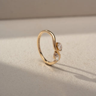 Angled view of a bypass diamond ring in 14 KT Yellow Gold, featuring a round and a pear-shaped lab-grown diamond embraced by flowing sculpted gold curves in an elegant open design, displayed against a beige backdrop.
