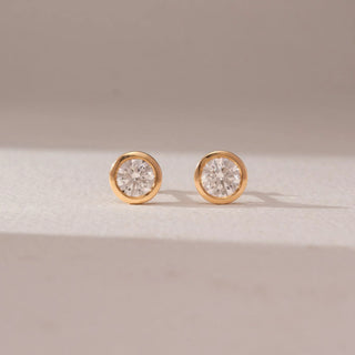 Front view of diamond stud earrings in 14KT yellow gold, featuring round brilliant diamonds in sleek bezel settings, displayed on a beige surface highlighting clean design and timeless sparkle.

