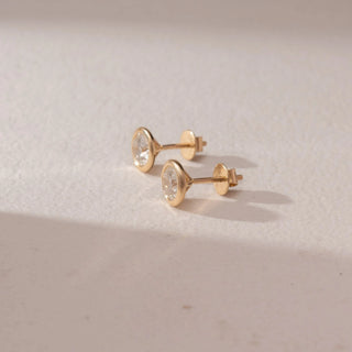 Side view of diamond stud earrings in 14KT yellow gold, featuring round brilliant diamonds in bezel settings with screw backs, placed on a beige surface highlighting smooth finish and subtle sparkle.

