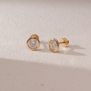 Angled view of diamond stud earrings in 14KT yellow gold, featuring round brilliant diamonds in sleek bezel settings with screw backs, set on a beige surface highlighting clean design and soft sparkle.
