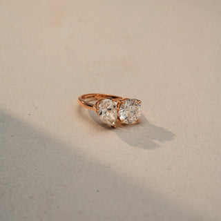 Side view of a toi et moi ring in 14 KT Rose Gold, featuring pear-shaped and round lab-grown diamonds set side by side in a modern two-stone design, displayed against a beige backdrop.