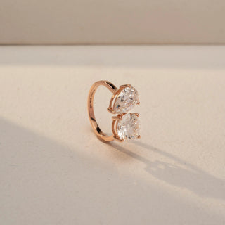 Angled view of a toi et moi ring in 14 KT Rose Gold, featuring pear-shaped and round lab-grown diamonds set side by side in a modern two-stone design, displayed against a beige backdrop.