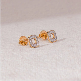 Angled view of square halo stud earrings in yellow gold, featuring an open centre framed by round lab-grown diamonds. Placed on a textured beige surface, the design casts a soft shadow.

