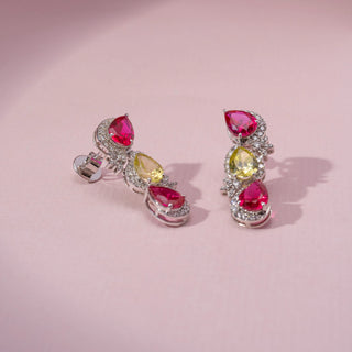 Angled view of ruby, yellow sapphire and lab-grown  round cut diamond drop earrings in 14 KT yellow gold, featuring pear-shaped ruby and yellow sapphire gemstones framed by pavé-set diamond halos with floral diamond accents.