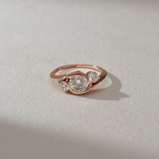 Top view of three-stone diamond ring in rose gold, featuring a round lab-grown diamond at the centre with two smaller round side stones in a flowing curved setting. Set on a soft neutral surface, it reflects modern elegance.