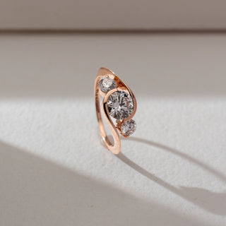 Side angled view of three-stone diamond ring in rose gold, featuring a round lab-grown diamond at the centre with two smaller round side stones in a flowing curved setting. Set on a soft neutral backdrop, it highlights elegance.
