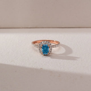 Top view of a halo ring featuring a cushion-cut blue lab-grown diamond centre stone, encircled by round accent diamonds and set on a pavé diamond band. Captured against a beige backdrop, the elegant halo design highlights vivid brilliance and sparkle.