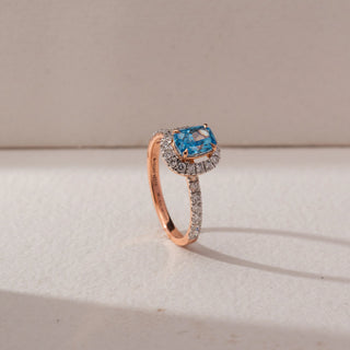 Angled side view of a halo ring featuring a cushion-cut blue lab-grown diamond centre stone, encircled by round accent diamonds and set on a pavé diamond band. Captured against a beige backdrop, the elevated halo design highlights brilliance and depth.