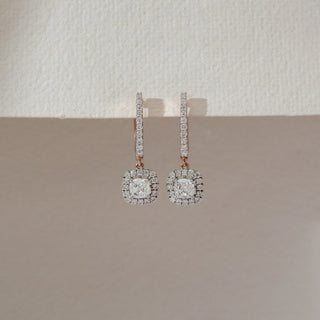 Front view of drop diamond earrings in rose gold, featuring a square halo lab-grown diamond pendant suspended from a vertical diamond-studded bar. Set against a soft beige textured background.