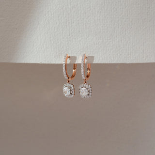 Angled view of rose gold hoop drop earrings with diamond-studded front, featuring a square halo lab-grown diamond pendant. Displayed against a soft beige textured background with gentle shadows.