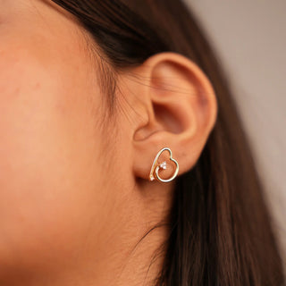 The image features side view of a female 14 KT yellow gold Stud Earring secured through a bombay screw weighing 2.187 grams, set with a 0.08 carat lab-grown diamond and displayed modelled on an earlobe. This earring boasts a contemporary heart-shaped design with clean, geometric edges, crafted in sleek yellow gold