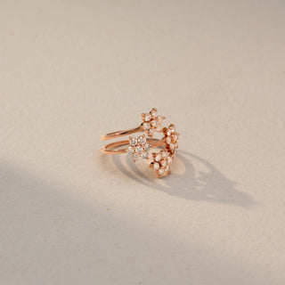 Side view of a floral wrap ring in 14 KT Rose Gold, featuring round lab-grown diamonds arranged in sparkling flower clusters along an elegant open spiral band, displayed against a beige backdrop.
