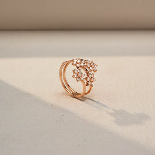 Angled view of a floral wrap ring in 14 KT Rose Gold, featuring round lab-grown diamonds arranged in sparkling flower clusters along an elegant open spiral band, displayed against a beige backdrop.