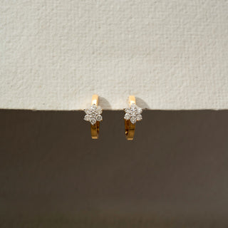 Front view of floral diamond hoop earrings in 18 KT Yellow Gold, featuring sparkling flower-shaped diamond clusters set on sleek gold hoops against a neutral backdrop.
