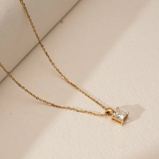 Angled view of yellow gold pendant featuring a radiant-cut lab grown diamond solitaire in a minimal open setting, suspended on a delicate chain against a soft beige surface.