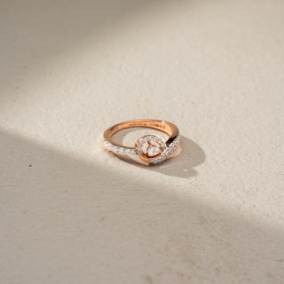 Top view of a swirl diamond ring in 14 KT Rose Gold, featuring a floating round lab-grown diamond framed by intertwining pavé-set diamond bands in an elegant crossover design, displayed against a beige backdrop.