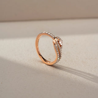 Angled view of a swirl diamond ring in 14 KT Rose Gold, featuring a floating round lab-grown diamond framed by intertwining pavé-set diamond bands in an elegant crossover design, displayed against a beige backdrop.