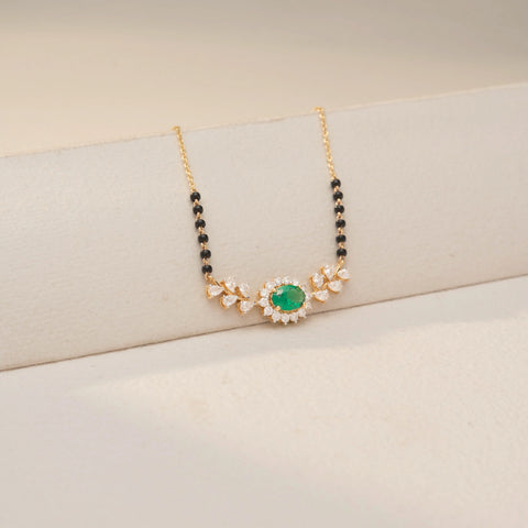 Close up view of an Emerald Diamond Mangalsutra in 14KT Dual Gold, featuring an oval emerald halo pendant with diamond leaf accents on a black bead chain. Captured against a beige backdrop.