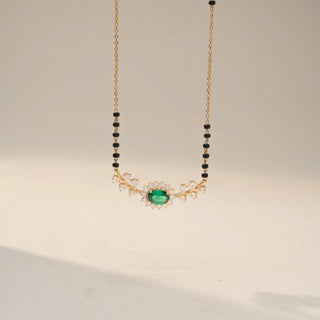 Front view of an Emerald Diamond Mangalsutra in 14KT Dual  Gold, featuring an oval emerald halo pendant with diamond leaf accents on a black bead chain. Captured against a beige backdrop.