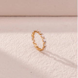 Side view of baguette diamond band in yellow gold, featuring vertically set lab-grown diamonds in a continuous zigzag pattern. Placed on a textured beige surface, the design casts a soft elongated shadow.
